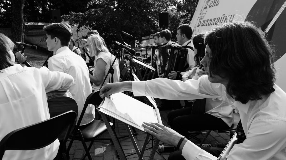 Outdoor orchestral performance with musicians playing various instruments in black and white.